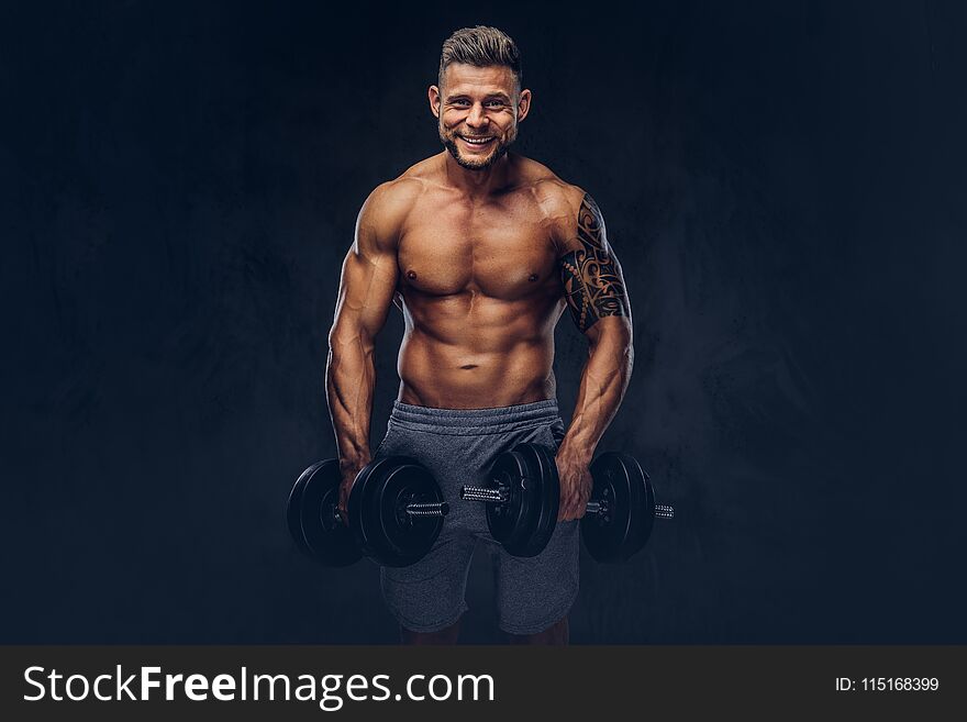 Smiling stylish bodybuilder with a tattoo on his arm, doing the exercises with dumbbells. Isolated on a dark background. Smiling stylish bodybuilder with a tattoo on his arm, doing the exercises with dumbbells. Isolated on a dark background.