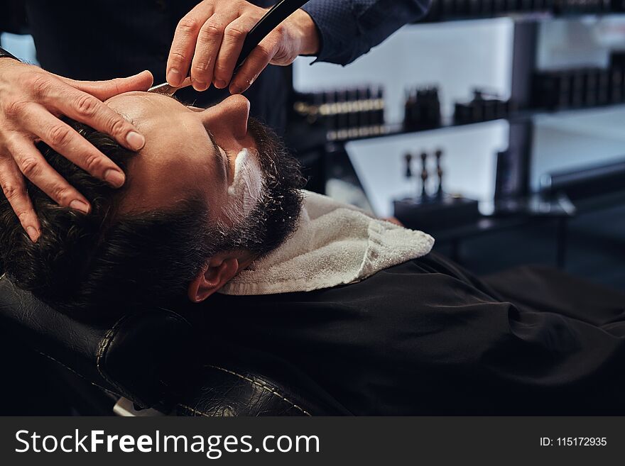 A handsome hipster bearded male sitting in an armchair in a barber shop while hairdresser shaves his beard with a dangerous razor.