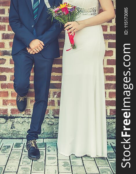 Bride and groom together in front of brick wall. Elegant bride in a white long wedding dress holding wedding bouquet of colorful flowers. Groom with crossed, folded arms leaning on the wall.