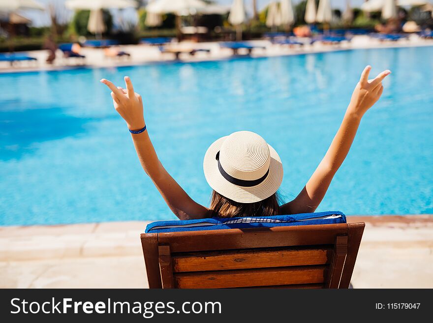 Rear view of Young beauty Woman in straw hat relaxing with hands up on chaise-lounge by the pool