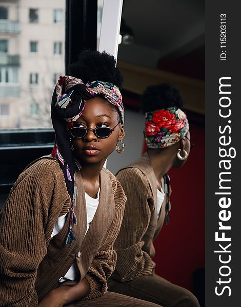 Woman Wearing White Shirt and Brown Coat With Sunglasses and Floral Turban Taking Selfie With Mirror Beside Her