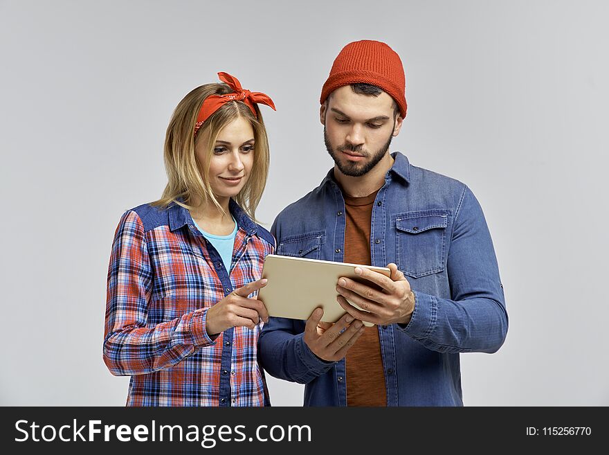 Young couple with attractive looks in casual hipster clothes look attentively at the tablet and learn something. Studio portrait on isolated background. Plaid shirts and hats