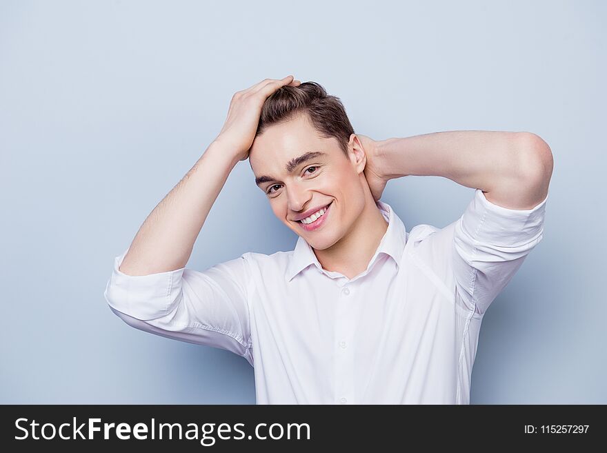 Portrait of happy with beaming smile young guy dressed in white shirt with rolled sleeves posing against gray background and touching his hair