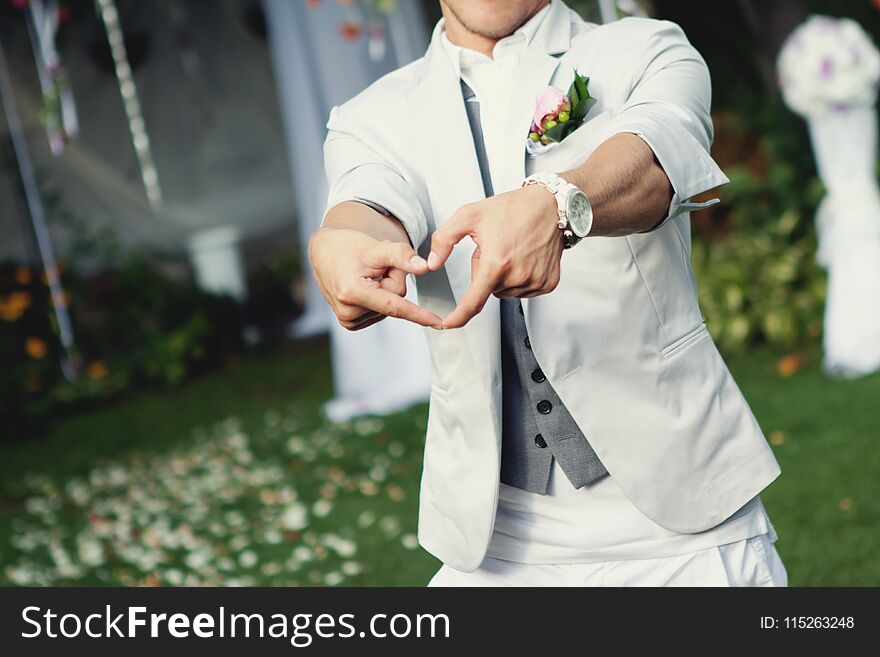 Happy stylish groom making a heart shape with hands. Groom in a bright suit with a white clock