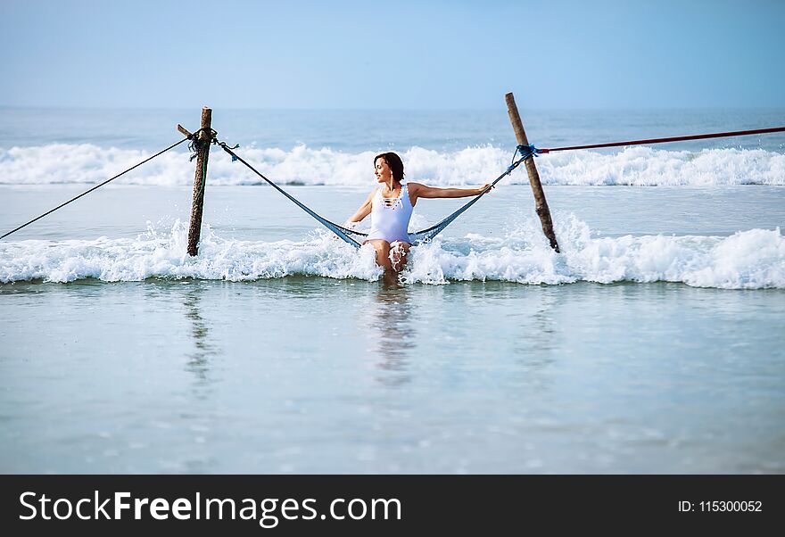Woman in white swimsuit sits in hammock swing over the ocean surf line