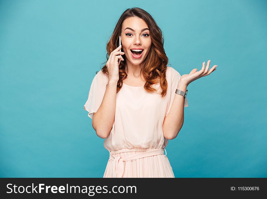 Portrait of a happy beautiful girl wearing dress talking on mobile phone and celebrating isolated over blue background