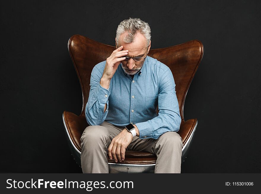 Image of intelligent gentleman 50s having grey hair and beard in glasses sitting on businesslike armchair with face downward isolated over black background