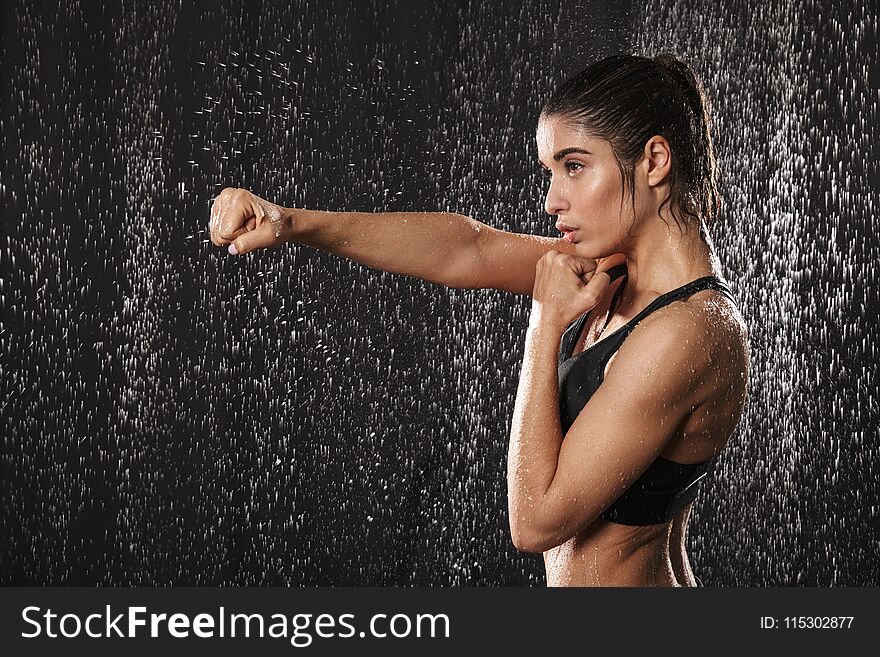 Photo in profile of focused athletic woman wearing black sportive bra punchning with clenched fist isolated over rain drops background