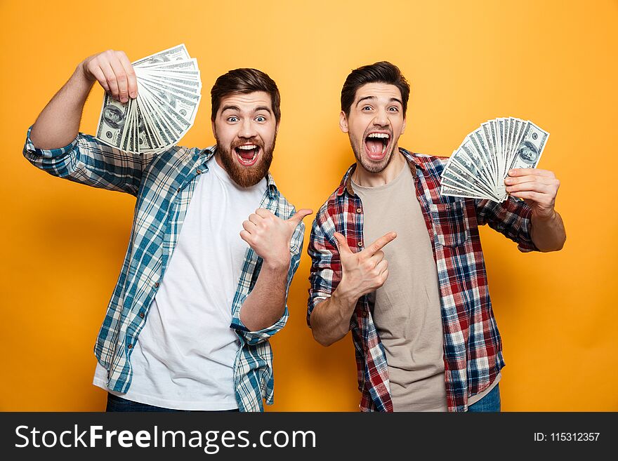 Portrait of a two excited oung men showing money banknotes and pointing fingers isolated over yellow background