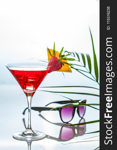 Colorful tropical cocktail in a martini glass decorated by slices of tropical fruits and a leaves on a reflecting table with sunglasses and palm tree leaves ona side. Negative space.