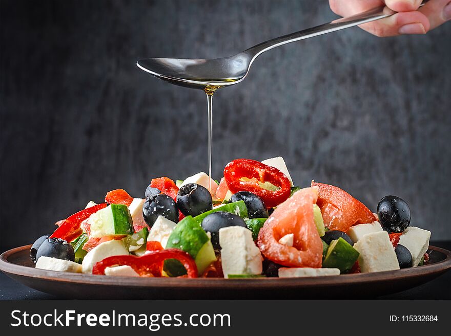 Woman`s hand is pouring a Greek salad with olive oil from a spoon on dark background. Woman`s hand is pouring a Greek salad with olive oil from a spoon on dark background