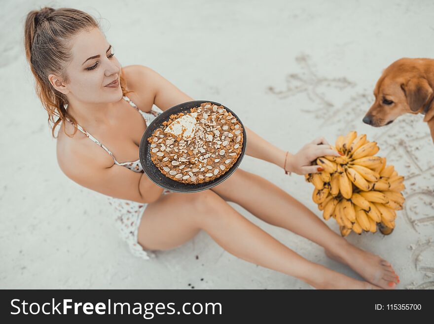 Woman siting on the sand with banana cake and bunch of bananas. Dog waiting for one piece.