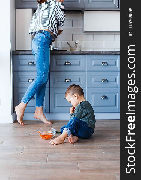 A Cute Little Boy Is Sitting On The Kitchen Floor With His Mother In The Background