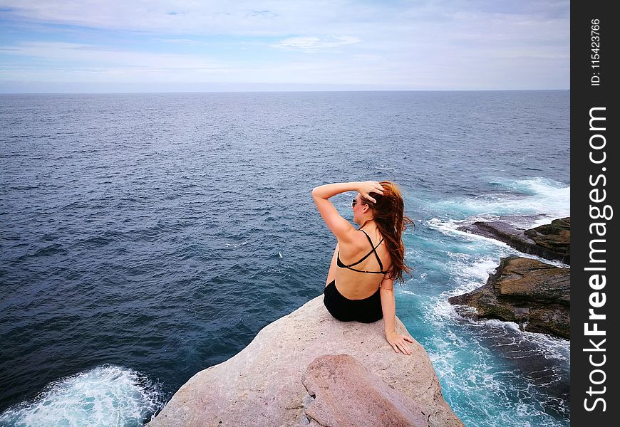 Woman in Black Swimwear Set Sitting on Top of Gray Rocky Cliff Facing Body of Water