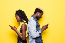 Phone Addiction. Portrait Of A Young Afro American Couple Standing Back To Back Using Mobile Phones Isolated Over Yellow Backgroun Stock Photo Phone Addiction. Portrait Of A Young Afro American Couple Standing Back To Back Using Mobile Phones Isolated Over Yellow Backgroun Stock Photo