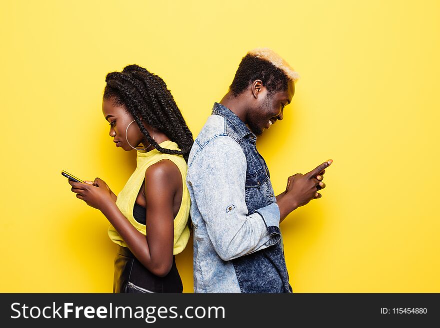 Phone addiction. Portrait of a young afro american couple standing back to back using mobile phones isolated over yellow backgroun