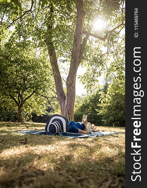 Woman lying relaxing in her back garden under a shady tree with a cold drink and wide brimmed sunhat in a low angle view over the lawn.