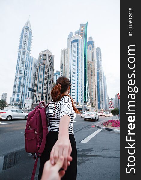 A beautiful girl with a backpack leads behind her and holds her hand against the background of tall buildings