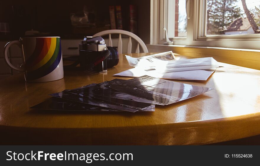 This vintage look photograph is warm with morning sun coming in the window. An old SLR camera from the 70`s sits next to a rainbow coffee mug. Black & white photographic prints are scattered across the table, along with the envelope from the processing lab & the pack of 35mm film negatives. This vintage look photograph is warm with morning sun coming in the window. An old SLR camera from the 70`s sits next to a rainbow coffee mug. Black & white photographic prints are scattered across the table, along with the envelope from the processing lab & the pack of 35mm film negatives.