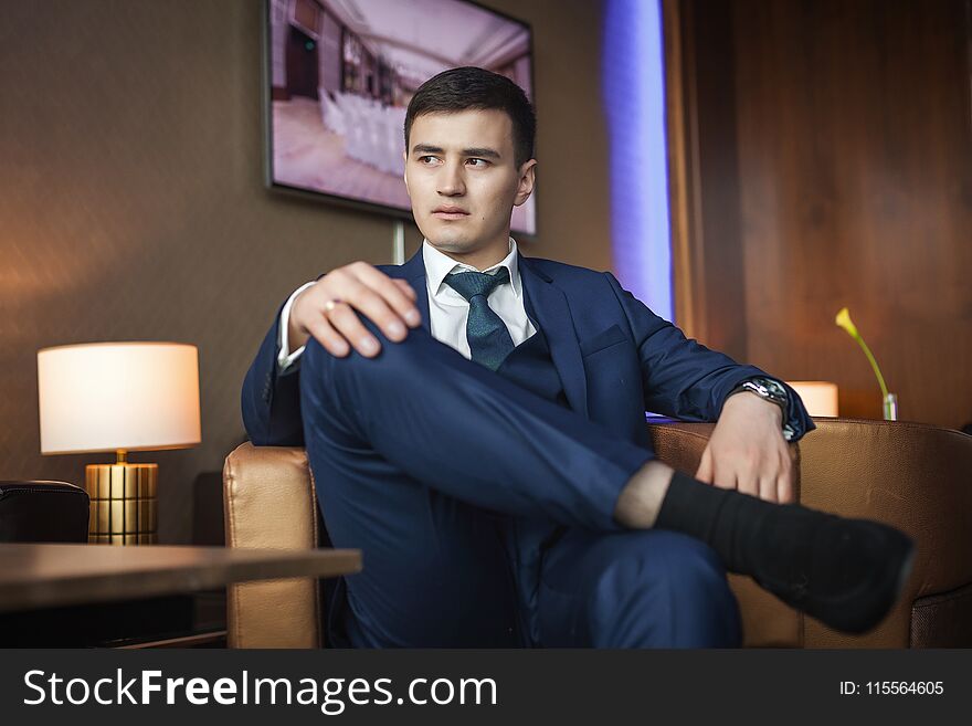 Young asian groom in dark blue suit sitting on a leather chair in the room of hotel and waiting for his bride. concept of a strong successful man. Young asian groom in dark blue suit sitting on a leather chair in the room of hotel and waiting for his bride. concept of a strong successful man