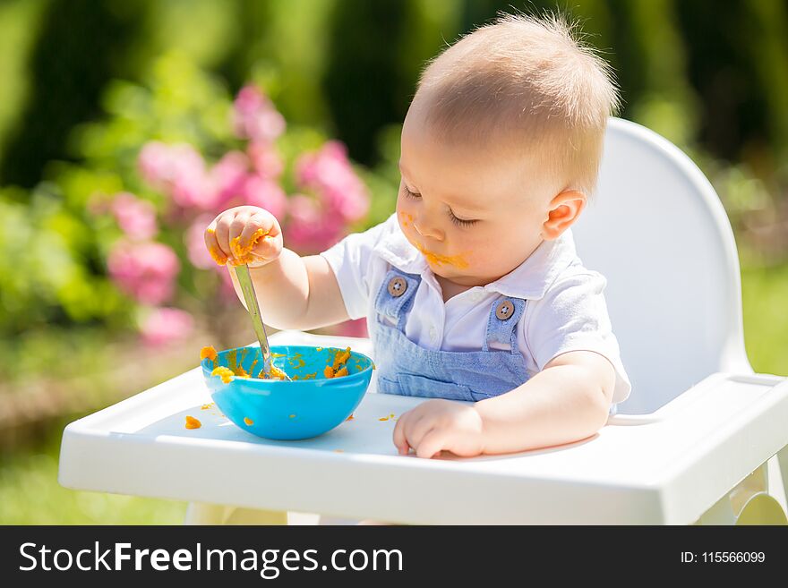 Cute toddler with messy face eating lunch in garden, sitting in baby chair. Outdoor shot.