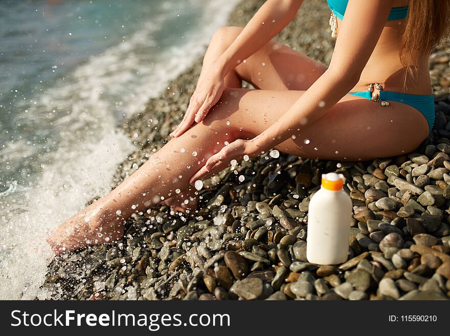 Slim woman tanned lower body in shape lying on pebble beach near sea waves and surf with sunblock cream bottle. Girl