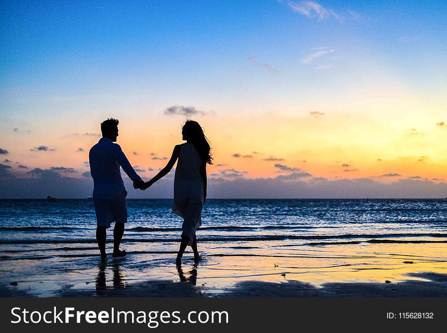 Man And Woman Holding Hands Walking On Seashore During Sunrise