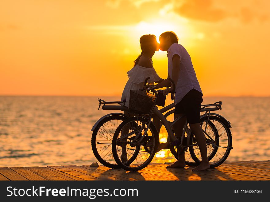 Photography of Man Wearing White T-shirt Kissing a Woman While Holding Bicycle on River Dock during Sunset