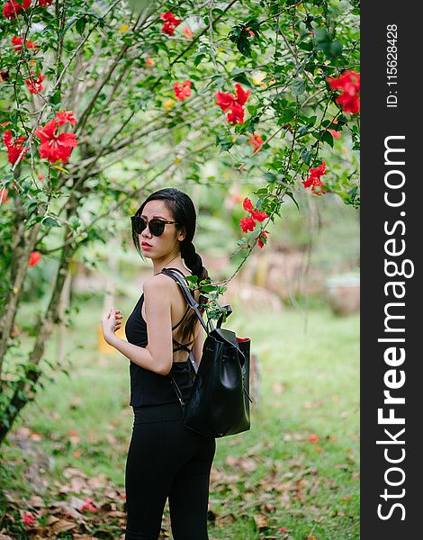 Woman Looking over Her Shoulder Wearing Black Sleeveless Top and Black Jeans Under a Red Hibiscus Plant at Daytime