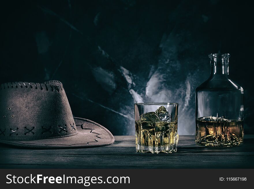 Glass of whiske and decanter with cowboy hat on wooden table. Toning.