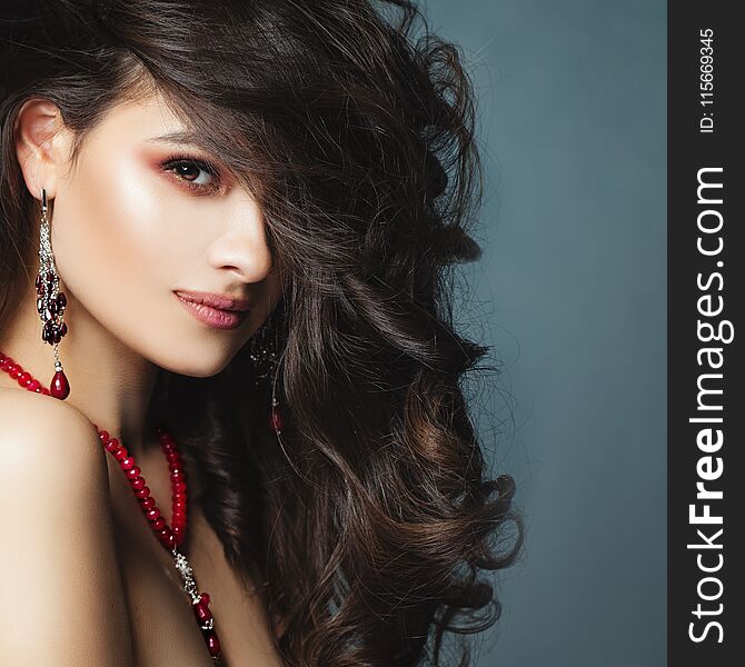 Perfect Brunette Woman Closeup Portrait. Young Female Face on Blue Background. Long Healthy Wavy Hair, Red Jewelry Necklace and Earrings