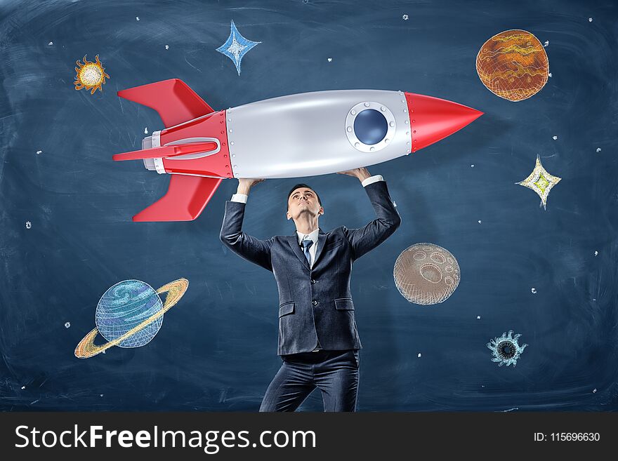 A businessman holds a model of a retro rocked above his head near drawings of planets and stars. Bring dreams to live. Business goals. Strength for success.