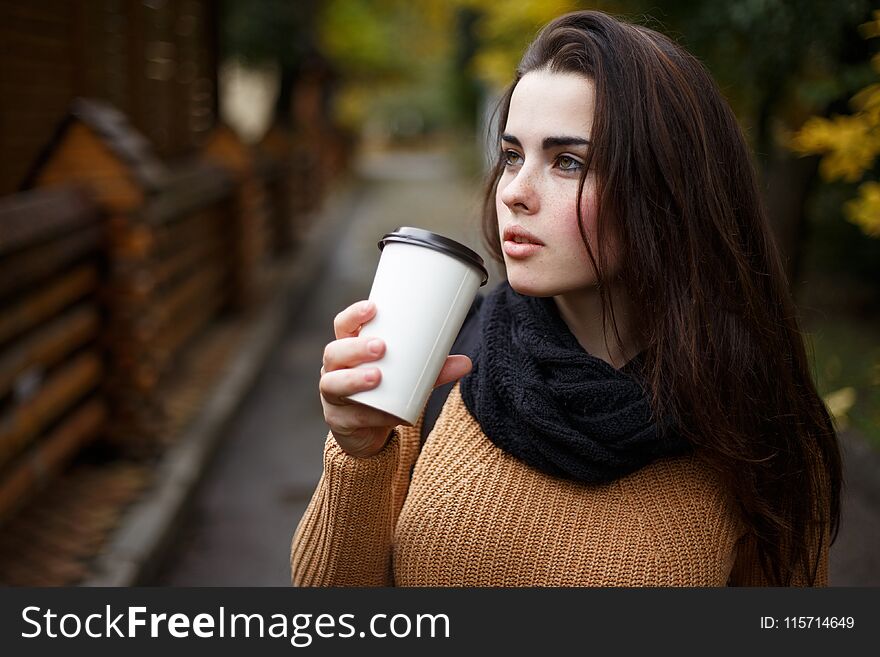 Young woman wearing knitted sweater walking in the autumn park and drinking take away coffee in paper cup. Breakfast on the go.