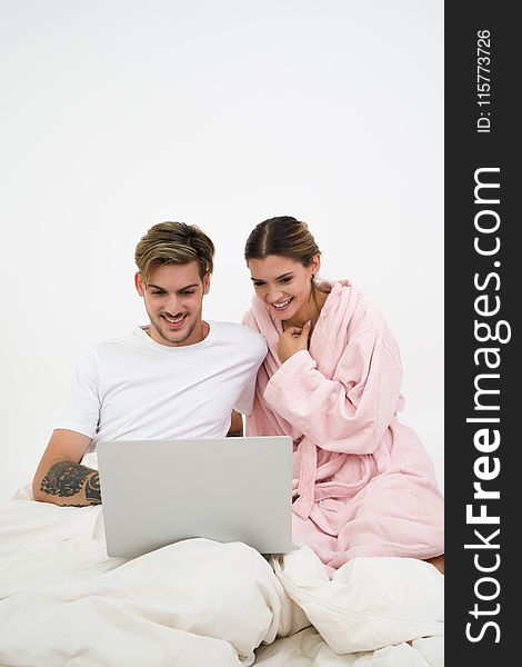 Man in White Crew-neck Shirt Sitting on Bed Beside Woman in Pink Bathrobe Looking at Laptop Computer