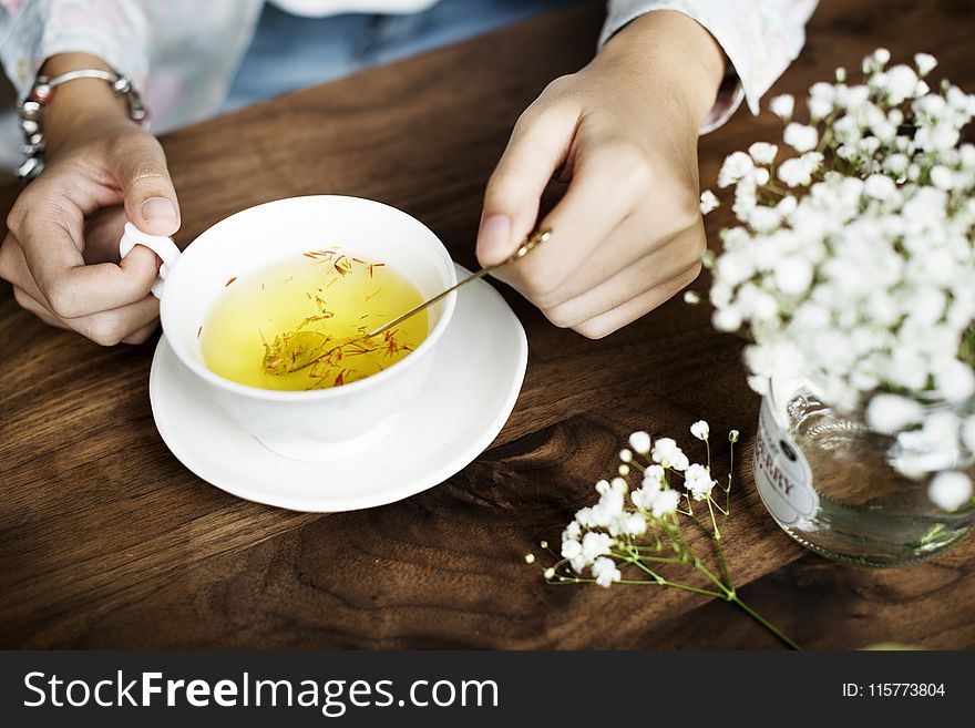 Person Holding Silver Spoon on White Ceramic Teacup With Yellow Liquid Inside