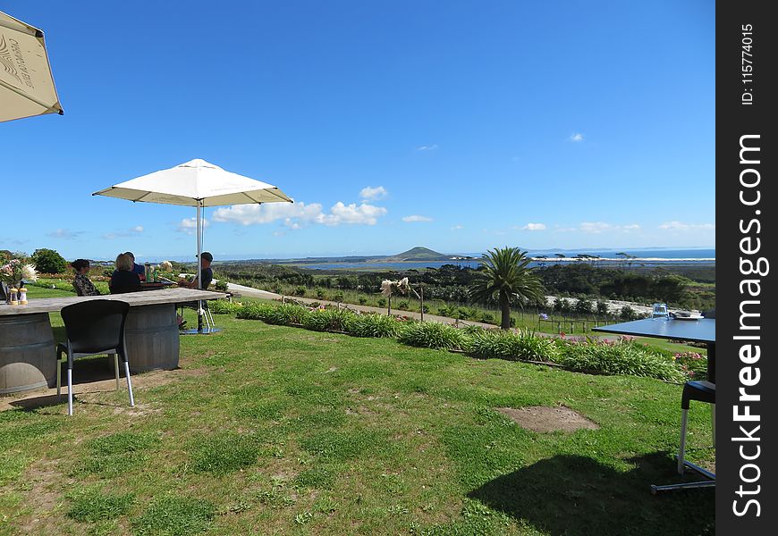 People Sitting In Front Of Patio Table With Parasol Placed On Hillside With View Of Body Of Water And Mountain