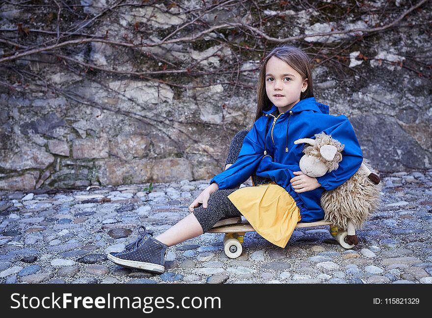 Cute little girl sitting on skateboard and holding toy ship on grey rock background. Cute little girl sitting on skateboard and holding toy ship on grey rock background.