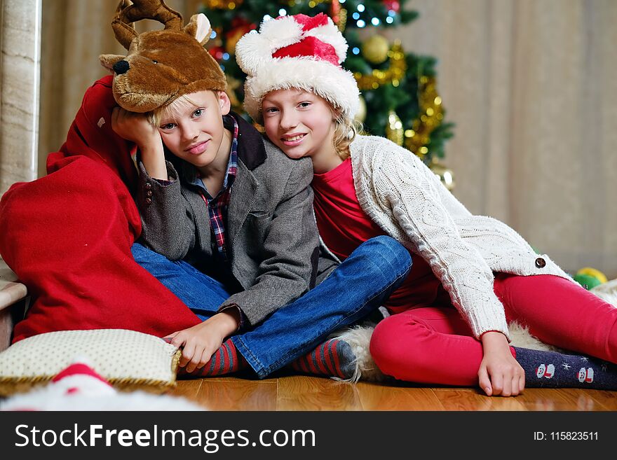 Smiling teenage girl and boy sitting on the floor with a Christmas tree in background.