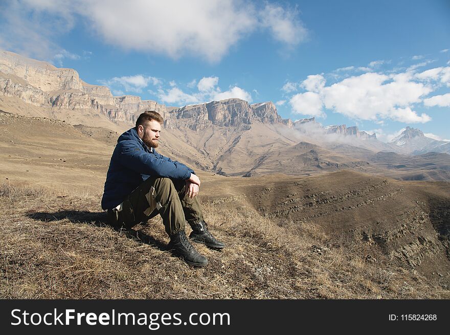 A portrait of a stylish bearded hipster sitting on a rock against the backdrop of epic rocks and contemplating into the distance thinking about life. The concept of tranquility and solitude with nature.