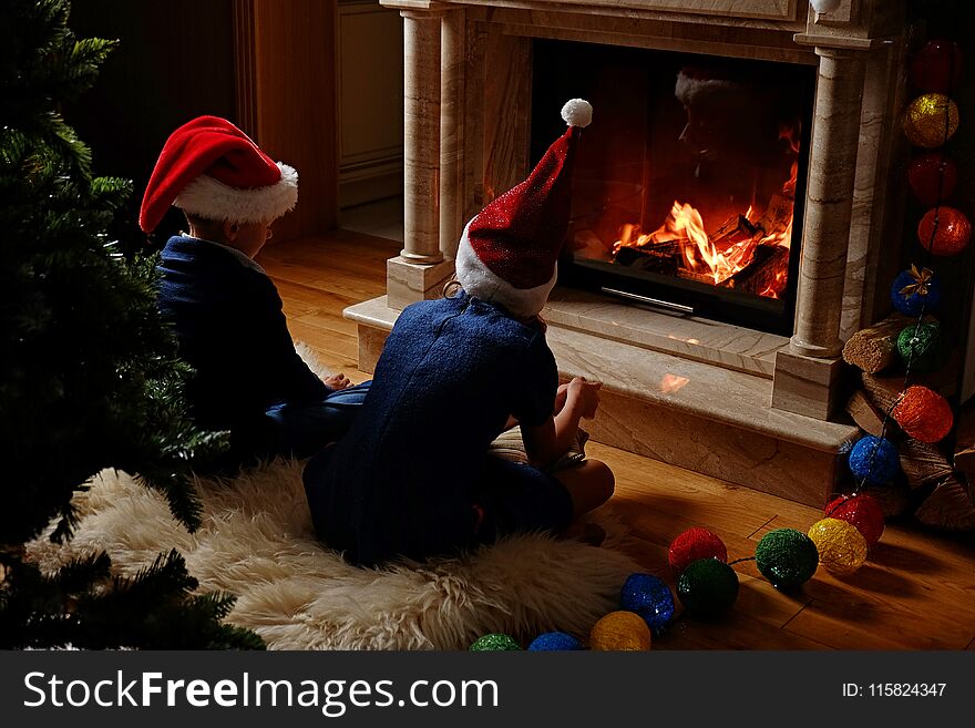 Two Cute Kids Sitting Near The Fireplace In Christmas Decorated Room.