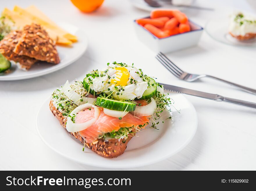Cereal bun with egg benedict, smoked salmon, sprout micro greens, onion and cucumber slices, cream cheese on the served white wooden table. Healthy breakfast concept. Selective focus