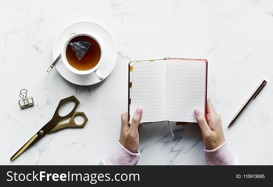 Person Holding Empty Book Near Pen and Shears With a Cup of Tea