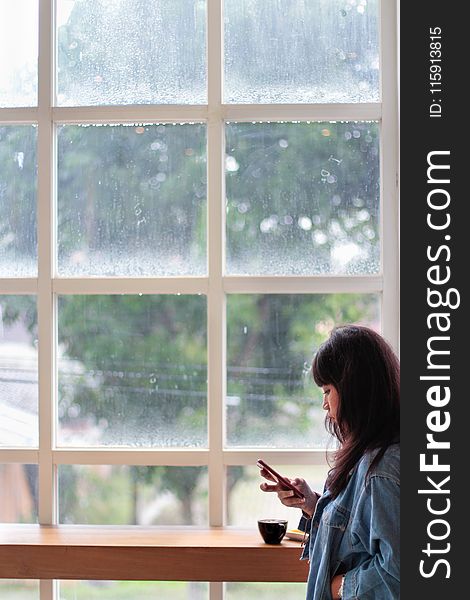 Woman Wearing Blue Denim Jacket Holding Smartphone Standing Beside Clear Glass Window