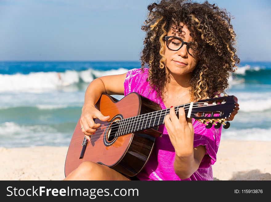 Woman Wearing Purple Shirt Playing Brown Classical Guitar While Sitting Near Shoreline With Water Splashing Background In Daytime