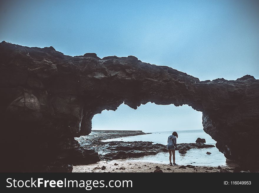 Person Standing On Beach Under Rock