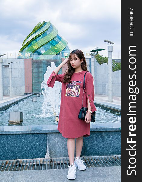 Woman Wearing Maroon Crew-neck Long-sleeved Dress Standing Next to Fountain Under Cloudy Sky