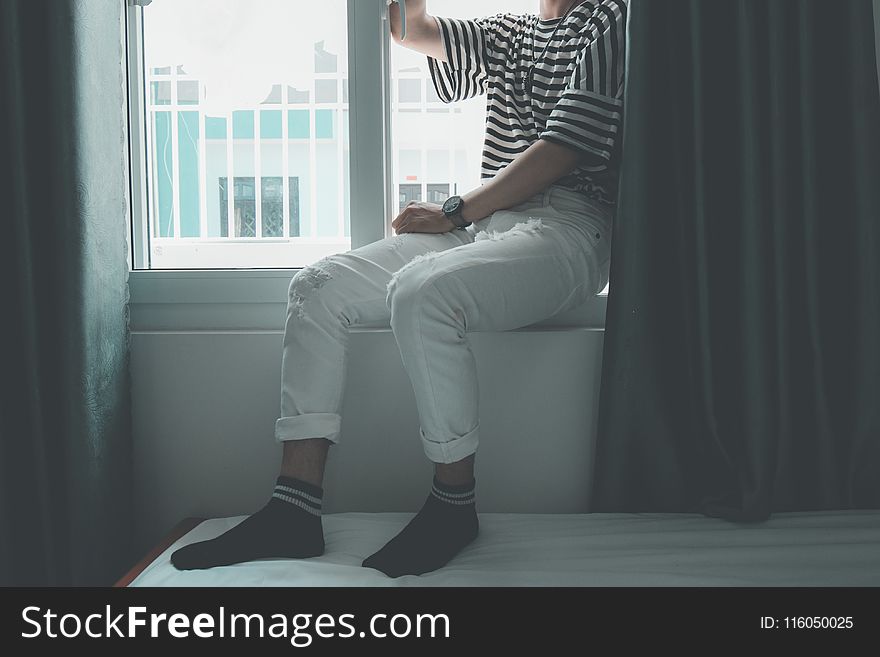 Woman Sitting on Window Stepping on White Bed Inside Well Lighted Room
