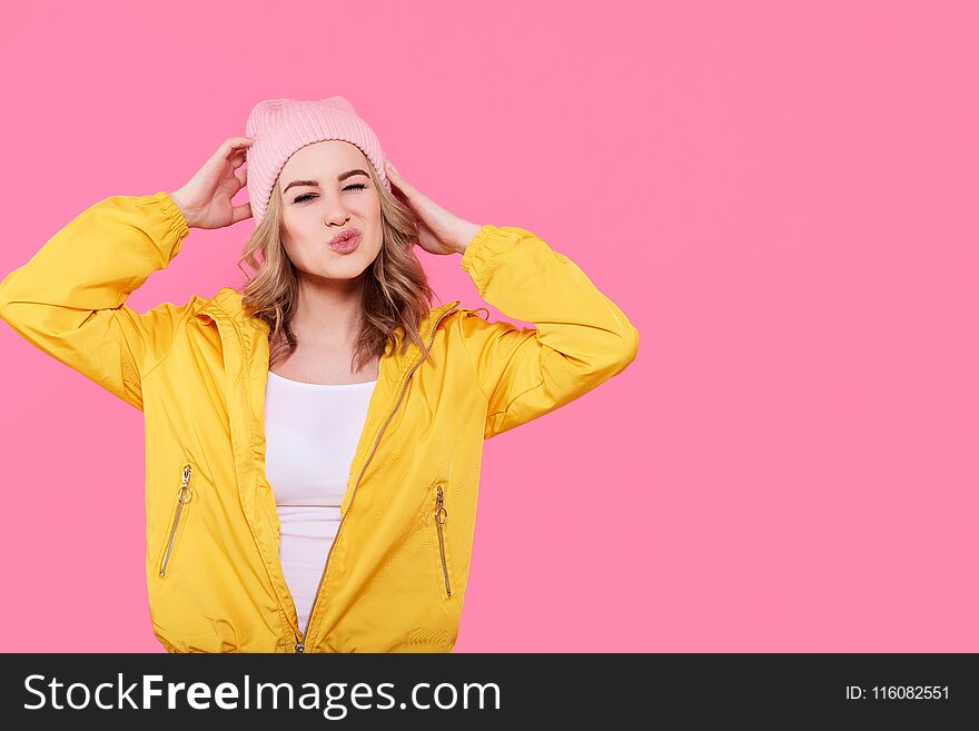 Crazy beautiful trendy girl in bright yellow jacket and pink beanie hat puckering lips. Cool young woman fashion portrait.