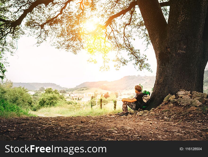 Little backpacker traveler rests under big tree on country road