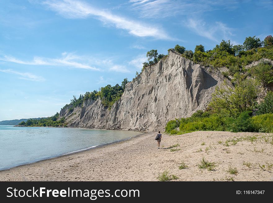 Woman Walks on Brown Seashore Near Cliff With Green Trees Under Blue and White Sky